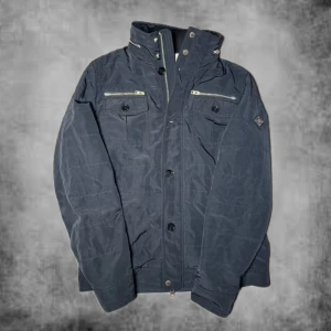 Mörkblå jacka från J.Lindeberg - Snygg mörkblå jacka från J.Lindeberg med hög krage, dragkedja och knappar framtill. Jackan har två bröstfickor med dragkedja och knappar samt diskret logga på ärmen. Perfekt för dig som gillar stilren och sportig look. Storlek XL passar mer som L