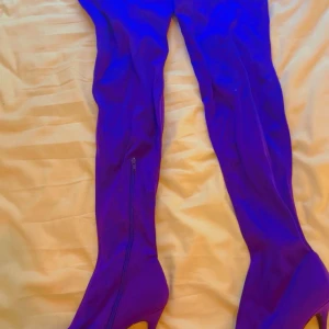 Lila overknee stövlar med klack - Säljer ett par snygga lila overknee stövlar med smal klack och dragkedja på insidan. Stövlarna har spetsig tå och är tillverkade i ett stretchigt syntetmaterial som sitter tight längs benet. Perfekta för att sticka ut i mängden.