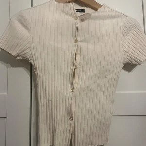 Ribbad kortärmad topp från Gina Tricot - Säljer en ribbad topp från Gina Tricot i färgen krämbeige. Toppen har korta ärmar, rund halsringning och knäppning framtill med små pärlemorknappar. Materialet är mjukt och stretchigt, perfekt för en clean och stilren look.