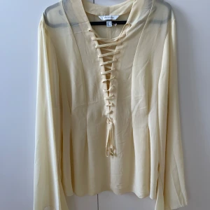 Beige blus med snörning från H&M Edition - Säljer en beige blus från H&M Edition med lång ärm och snygg snörning framtill. Blusen är tillverkad i ett tunt, lätt material som ger en flowy känsla. Perfekt för dig som gillar stilrena detaljer och en chic look.