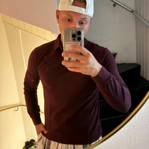 Vinröd pikétröja från Polo Ralph Lauren - Säljer en vinröd långärmad pikétröja från Polo Ralph Lauren med klassisk krage och knappar framtill. Tröjan har den ikoniska blå loggan broderad på bröstet och är tillverkad i mjuk bomull. Perfekt för dig som gillar stilren och tidlös design.