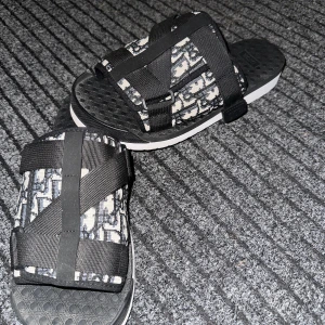 Svarta Dior sandaler - Säljer ett par nya svarta sandaler från Dior med ikoniskt monogrammönster i vitt och svart. De har breda, justerbara remmar i textil och en bekväm, mönstrad sula med Dior-logga. Perfekta för sommaren och ger en lyxig touch till din outfit., Används tre-fyra gånger, pris kan diskuteras!