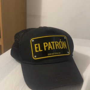 Svart keps John Hatter & Co El Patrón - Svart keps från John Hatter & Co med mesh på sidorna och justerbar snapback. Framtill sitter en svart och gul skylt med texten 'EL PATRÓN'. Kepsen har en cool streetstyle-vibe och är perfekt för dig som vill sticka ut.