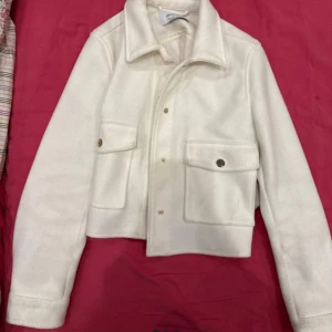 Vit overshirt från Stradivarius - Snygg vit overshirt från Stradivarius med stora fickor framtill och guldiga knappar. Jackan har klassisk krage, raka linjer och är tillverkad i ett mjukt material som känns lite ulligt. Perfekt för lager-på-lager och enkel att styla till allt.