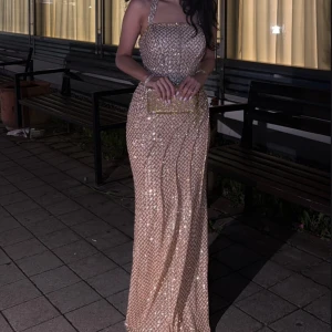 Champagnefärgad glittrig långklänning - Supersnygg långklänning i champagnefärg med glittriga paljetter och rutigt mönster. Klänningen har halterneck, markerad midja och figurnära passform. Materialet är syntet med insydda detaljer och dragkedja i ryggen. Perfekt för bal eller nyår!