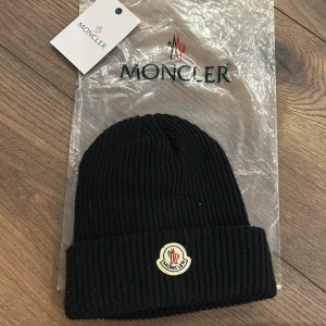 Svart mössa från Moncler - Svart ribbstickad mössa från Moncler med klassisk uppvikt kant och broderad logotyp framtill. Perfekt accessoar för kyliga dagar med stilren design och exklusiv känsla.