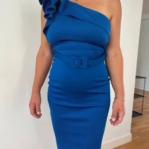 Kornblå one shoulder klänning med volang - Säljer en kornblå, figurnära one shoulder-klänning med volangdetalj över axeln och matchande bälte i midjan. Klänningen har en elegant, slät yta och är axelbandslös på ena sidan. Perfekt för dig som vill sticka ut med en färgstark look.