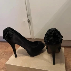 Svarta pumps med paljettdetalj - Snygga svarta pumps från Bershka i satinliknande material med öppen tå och hög klack. Baktill är skorna dekorerade med svarta paljetter som ger extra glans. Perfekta för dig som vill sticka ut med en elegant och trendig look. Klacken är 13cm