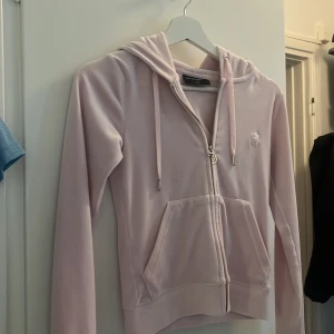 Ljusrosa velourhoodie Juicy Couture - Superfin ljusrosa hoodie från Juicy Couture i mjuk velour. Tröjan har dragkedja med snyggt J-hänge, huva och fickor framtill. Perfekt för dig som gillar den klassiska Y2K-stilen och vill ha något riktigt cozy och trendigt.