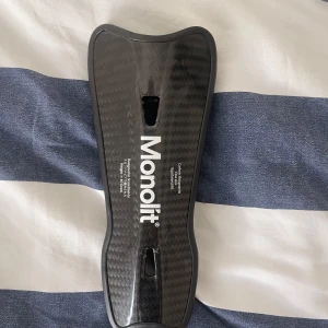 Monolit benskydd  - Säljer ett par svarta Monolit benskydd i kolfiber med snygg, sportig design. Lätta och smidiga med ergonomisk passform och vadderad insida för extra komfort. Perfekta för dig som vill ha maxad skyddsnivå på fotbollsplanen. De är lite skadade med små sprickor.