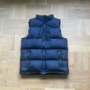 Mörkblå dunväst från Ralph Lauren - Mörkblå dunväst från Ralph Lauren med klassisk puffer-design, hög krage och dragkedja framtill. Västen har två fickor med dragkedja och en liten röd logga broderad på bröstet. Perfekt för lager-på-lager under kyliga dagar.