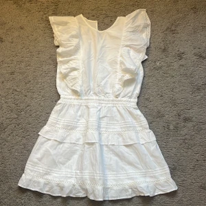 Vit volangklänning med spetsdetaljer - Superfin vit kortklänning med volanger på axlarna och kjolen, samt spetsdetaljer längs kanterna. Klänningen har resår i midjan för en snygg passform och är ärmlös. Perfekt för sommarens alla tillfällen!