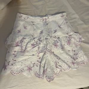 Broderade vita shorts från By Malina - Superfina vita shorts från By Malina med lila broderade blommor och volangdetaljer. De har hög midja, dragkedja i sidan och är gjorda i bomull. Perfekta för sommarens alla tillfällen när du vill sticka ut lite extra.