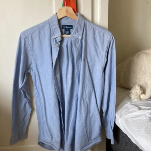 Blå skjorta från Ralph Lauren - Klassisk ljusblå skjorta från Ralph Lauren med broderad logga på bröstet. Skjortan har lång ärm, knappar framtill och button-down krage. Tillverkad i mjuk bomull som känns skön mot huden. Perfekt för en clean och stilren look. Skick 8/10, insidan av skjortan är lite sliten men inget man lägger märke till. Strlk: 16 år.