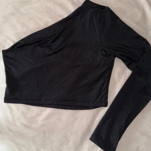 Svart oneshoulder topp med öppen rygg - Säljer en svart, tight och croppad topp med oneshoulder-design och lång ärm. Toppen har en öppen rygg som ger en cool och modern look. Perfekt för en trendig look!