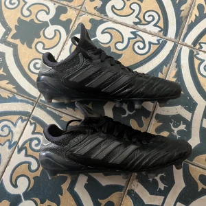 Adidas copa fotbollsskor - Säljer ett par helsvarta adidas fotbollsskor med klassisk design och tre ränder på sidan. Skorna har snörning, rund tå och fasta dobbar för gräsplan. Ovandelen är i skinn för bra känsla och passform. Perfekta för dig som vill ha stilrena och funktionella skor på planen.