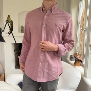 Rosa skjorta från Ralph Lauren M - Säljer en snygg rosa skjorta från Ralph Lauren i custom fit. Skjortan har klassisk button-down krage, långa ärmar och broderad logga på bröstet. Tillverkad i mjuk bomull och stängs med vita knappar framtill. Perfekt för en clean och stilren look.
