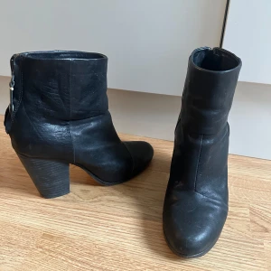Svarta stövletter i skinn med klack från Rag&Bone - Säljer ett par svarta stövletter i äkta skinn med kraftig klack och dragkedja bak. Skorna har en rund tå och stilren design som passar till många outfits. Perfekta för dig som gillar boots med lite höjd och snygg siluett. Klacken är 9cm