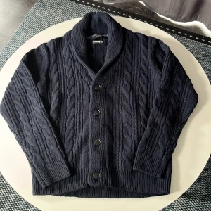 Mörkblå kabelstickad kofta Nano Universe - Kabelstickad mörkblå kofta från Nano Universe i cashmere-mix. Koftan har sjalkrage, stora knappar framtill och ribbade muddar. Perfekt för lager-på-lager och chill vibes. Materialmix med ull och kashmir för extra mjuk känsla.