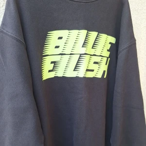 Svart Billie Eilish sweatshirt - Cool svart sweatshirt med stort neongult Billie Eilish-tryck på bröstet. Tröjan har en avslappnad passform och är perfekt för dig som vill sticka ut med en streetstyle-look. Mjuk och skön, passar perfekt till jeans eller joggers.