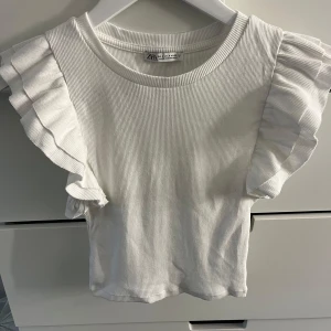 Vit ribbad topp med volangärmar Zara - Superfin vit ribbad topp från Zara med korta volangärmar. Toppen har rund halsringning och är tillverkad i mjuk bomull. Perfekt för dig som gillar en söt och stilren look med extra detalj på ärmarna.