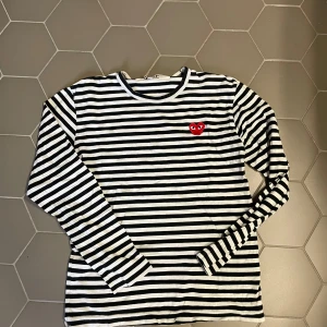 Randig långärmad tröja Comme des Garçons - Svartvit randig långärmad tröja från Comme des Garçons Play med det ikoniska röda hjärtat broderat på bröstet. Tröjan är i mjuk bomull och har en klassisk rund halsringning. Perfekt för dig som gillar stilrena och trendiga plagg.