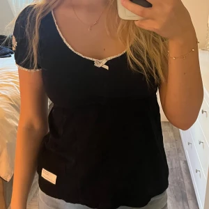 Svart Odd Molly topp med spetsdetaljer - Supersöt svart topp från Odd Molly, modell Fella Top. Toppen har korta ärmar, vit spetskant runt halsringning och ärmslut samt en liten rosett framtill. På ena ärmen finns ett broderat Odd Molly-märke. Perfekt för dig som gillar feminina detaljer.