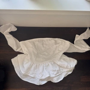 Vit broderad tubtopp med volang - Supersöt vit tubtopp med broderade blommor och volangkant. Toppen har breda knytband som kan knytas i ryggen för en snygg detalj. Perfekt för dig som gillar romantisk och feminin stil. Materialet känns lite tjockare och har struktur.