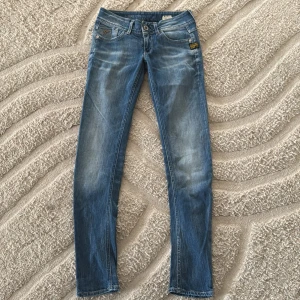 Blå skinny jeans från G-Star - Snygga blå jeans från G-Star med slitningar och klassisk femficksdesign. Modellen är skinny med låg midja och har coola detaljer som logga på fickan och metallknapp (tappat lite färg) framtill. Perfekta för dig som gillar en tajt passform och streetig look. W26 L30 