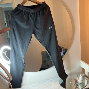 Svarta träningsbyxor från Under Armour - Svarta träningsbyxor från Under Armour med elastisk midja och logga på låret. Byxorna är i löst passform och tillverkade i ett lätt syntetmaterial, perfekta för träning. Modellen är enkel och stilren utan extra detaljer.