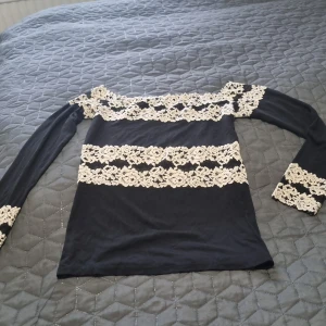 Svart offshoulder topp med spets från Intimissimi - Supersnygg svart offshoulder topp från Intimissimi med långa ärmar och breda dekorativa spetsband i vitt över bröst, midja och ärmslut. Blommig spets ger en romantisk vibe och toppen har en figurnära passform. Perfekt för dig som vill sticka ut med stil. Den är I jätte bra skick storlek S. 