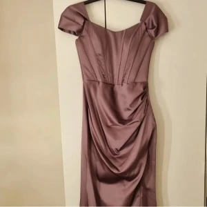 Ljuslila långklänning med snörning - Elegant ljuslila långklänning i satinliknande material med offshoulder-ärmar, markerad midja och snygg drapering framtill. Baksidan har dekorativ snörning som ger en extra detalj. Perfekt för dig som vill sticka ut med en romantisk och trendig look.