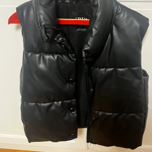 Svart dunväst i skinnimitation från Zara - Stilren svart dunväst från Zara i skinnimitation med quiltad design. Västen har hög krage, dragkedja och tryckknappar framtill. Perfekt för lager-på-lager och ger en edgy vibe till din outfit.
