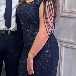 Mörkblå glittrig långklänning med pärldetaljer - Säljer en elegant mörkblå långklänning med glittriga paljetter och pärldetaljer längs axlarna som hänger ner i kedjor. Klänningen har figurnära passform och ett unikt mönster av paljetter över hela tyget. Perfekt för bal eller festliga tillfällen.