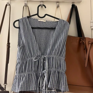 Blårutig ärmlös topp från Zara - Supersöt ärmlös topp från Zara i blåvitt rutigt mönster. Toppen har djup v-ringning, knytband i midjan och volang nedtill. Perfekt för sommarens alla tillfällen och ger en fräsch och trendig look. Köpt i somras, använt fåtal gånger men är lite smutsiga vid etiketten 💕