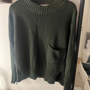Mörkgrön stickad tröja med ficka - Snygg mörkgrön stickad tröja med ribbad krage och nederkant. Tröjan har en liten ficka framtill och en cool patchdetalj på ena ärmen. Passar dig som gillar avslappnad stil och vill ha något som sticker ut lite extra.