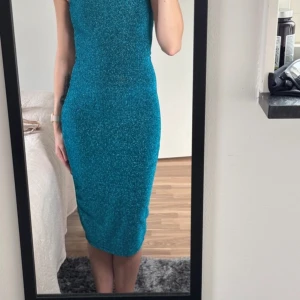 Glittrig blå midiklänning med slits - Supersnygg glittrig blå klänning med tight passform och slits bak. Klänningen är ärmlös och har en midilängd som sitter figurnära hela vägen. Perfekt för dig som vill sticka ut med lite extra glitter.