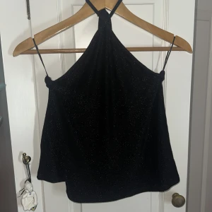 Svart glittrig halterneck topp - Snygg svart glittrig halterneck topp med smala axelband som korsas i nacken. Materialet är mjukt och har en skimrande finish som ger en extra touch. Toppen är ärmlös och har en loose passform, perfekt för att sticka ut med en cool vibe. Jag använt den till fester och utekvällr passar perfekt och är sååå snygg!! Den kommer bar inte till användning för mig längre ‼️🥰