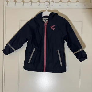 Lupilu - regnjacka, storlek 86/92 - Lupilu marinblå regnjacka storlek 86/92  Säljer även Lupilu regnbyxor i samma storlek, säljer båda tillsammans för ett bättre pris. Marinblå regnjacka med rosa detaljer. Jackan har en huva, dragkedja framtill och två fickor. Det finns reflexer på ärmarna och ett tryck med hjärtan på bröstet.  Skickar med frakt eller avhämtning i Växjö. Vid flera köp kan du som köpare välja samfrakt. Fraktpriset ingår inte i priset. 