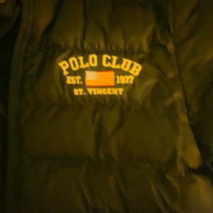 Svart pufferjacka från Polo Club, storlek M  - Svart pufferjacka från Polo Club med broderad logga och amerikansk flagga på bröstet. Jackan har huva, dragkedja framtill och två sidofickor. Perfekt för kalla dagar och har en sportig vibe med coola detaljer.