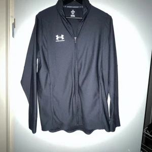 Svart zip k  kofta från under armor - Svart kofta från Under Armour med vit logga på bröstet. Tröjan har hel dragkedja framtill, ståkrage och långärmade ärmar. Storlek M men passar som L. Fick som present men lite stor