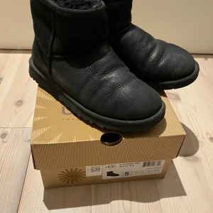 Svarta UGG Classic Mini boots storlek 36. - Mysiga svarta UGG Classic Mini boots med rund tå och platt sula. Skorna är fodrade med mjuk päls och har en klassisk siluett i mocka och skinn. Perfekta för kalla dagar när du vill hålla fötterna varma och bekväma. Innermått 23,3.