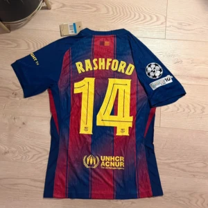 FC Barcelona Rashford tröja Nike M - Säljer en officiell FC Barcelona fotbollströja från Nike i storlek M med Rashford och nummer 14 på ryggen. Tröjan har klassiska blå och röda vertikala ränder, gula detaljer och Champions League-märke på ärmen. Materialet är lätt och andas, perfekt för match eller träning.