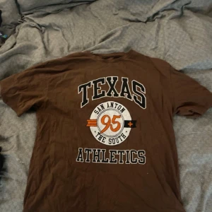 Brun Texas Athletics t-shirt XL - Brun t-shirt med tryck 'TEXAS ATHLETICS' och 'San Anton 95 The South' i vitt, svart och orange på framsidan. Klassisk rund hals och korta ärmar. Skön och avslappnad passform, perfekt för dig som gillar sportiga college-vibbar. Materialet är mjukt bomull.