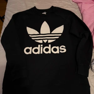 Svart Adidas sweatshirt med logga - Svart sweatshirt från Adidas med stor vit treklöver-logga och text på bröstet. Tröjan har rund hals och långa ärmar, tillverkad i mjuk bomullsmix som är skön att ha på sig. Perfekt för dig som gillar sportig och clean streetstyle.