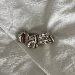 Silvrig hårklämma med vågig form - Unik hårklämma i blankt silvrigt metallmaterial med cool, skulptural och vågig design. Perfekt för att ge din frisyr en edgy touch och sticker verkligen ut med sin glansiga yta och moderna look. Klämman har ett stabilt spänne på baksidan för enkel användning.