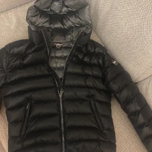 Svart Colmar pufferjacka med huva - Säljer en svart pufferjacka från Colmar med huva och två sidofickor med dragkedja. Jackan har ett stilrent, quiltat mönster och Colmar-logga på ärmen. Perfekt för dig som vill ha en snygg och varm jacka med modern look. 