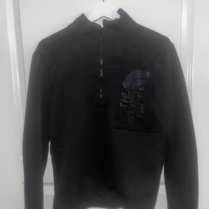 Svart half zip tröja The North Face - Svart långärmad tröja från The North Face med half zip och stor logga på bröstet. Tröjan har en hög krage och är gjord i mjukt syntetmaterial som är skönt mot huden. Perfekt för dig som gillar stilren och sportig look. Skick 8/10