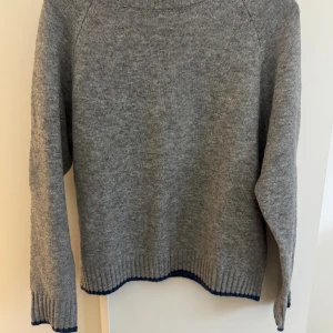 Grå stickad tröja med blå kant - Mysig grå stickad tröja med mörkblå ribbad kant vid hals, ärmslut och nederkant. Tröjan har en klassisk rund hals och långärmade, loose passform. Perfekt för dig som gillar enkel och stilren design med en liten twist.