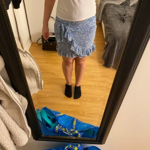 Blå blommig omlottkjol med volang - Supersöt blå kort omlottkjol med vitt blommönster och volangkant. Kjolen har knyt i sidan och en flowy känsla som ger en riktigt somrig vibe. Perfekt för dig som gillar romantiska detaljer och vill ha en kjol som sticker ut lite extra.